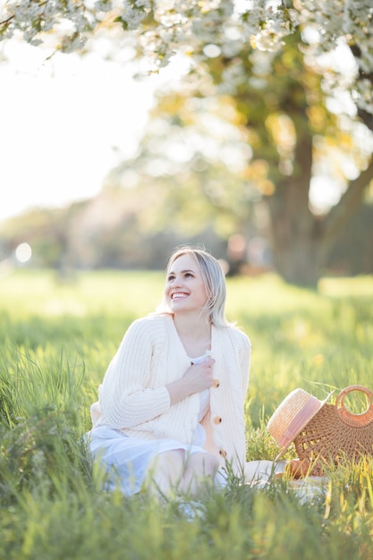 Beautiful young woman in a wicker hat is resting on a picnic in a blooming garden