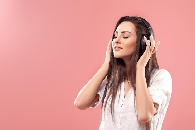 Beautiful young woman in wireless headphones listening to music and dancing on pink background