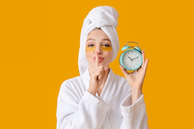 Beautiful young woman with alarm clock showing silence gesture on color surface