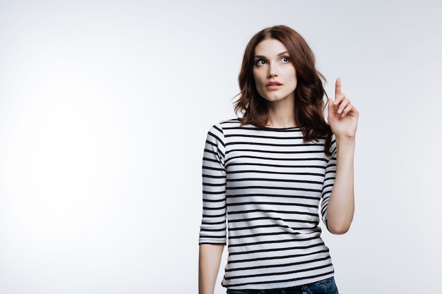 Beautiful young woman with an auburn hair raising her finger, having remembered to do something important on grey