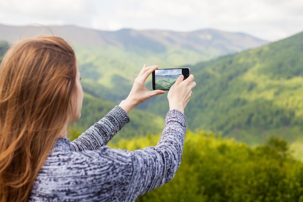 美しい髪の美しい若い女性は、緑の自然の彼女の黒い携帯電話で写真を撮っています