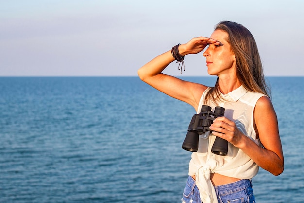 Beautiful young woman with binoculars on the cliff looks at the sea.