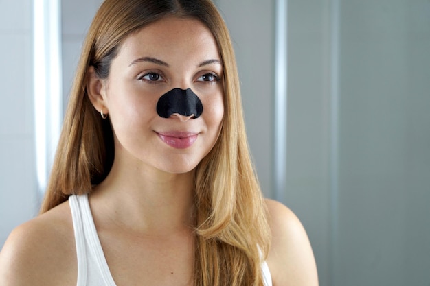 Beautiful young woman with black charcoal nose strip looking at mirror at home