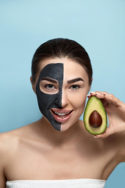 Красивая молодая женщина с черной маской из глины на лице Красивое лицо девушки держит половинку авокадо