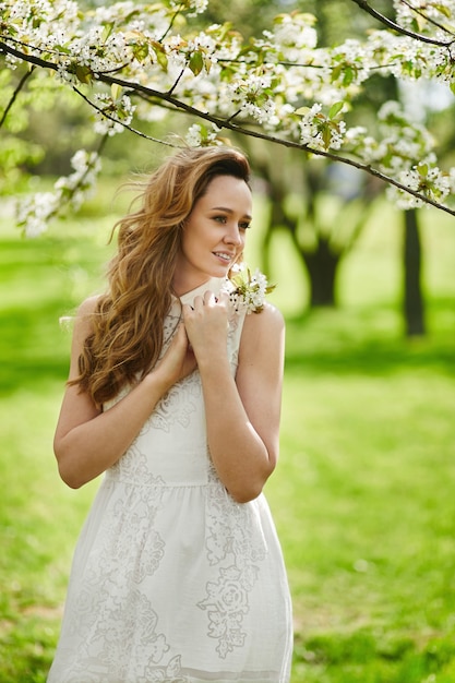 Beautiful young woman with blond hair in fashionable white dress posing near a flowering tree at a blooming garden at summer sunny day