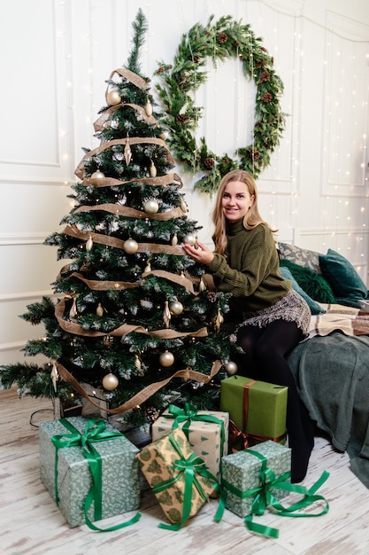 A beautiful young woman with blond hair sits near a Christmas tree and decorates with toys. New Years is soon. Christmas atmosphere in a cozy home