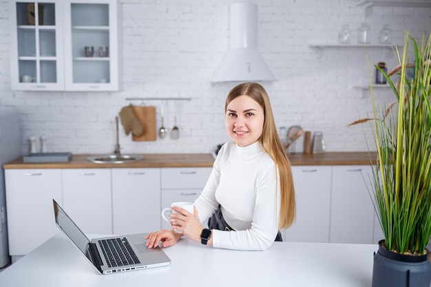 흰색 터틀넥 스웨터에 금발 머리를 한 아름다운 젊은 여성이 흰색 부엌에 서서 노트북에서 일하고 커피를 마십니다. 집에서 원격으로 작업