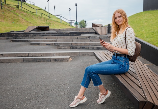 街の通りでスマートフォンでブロンドの髪のメッセージを持つ美しい若い女性。晴れた日に通りで電話でSMSを入力する女の子。