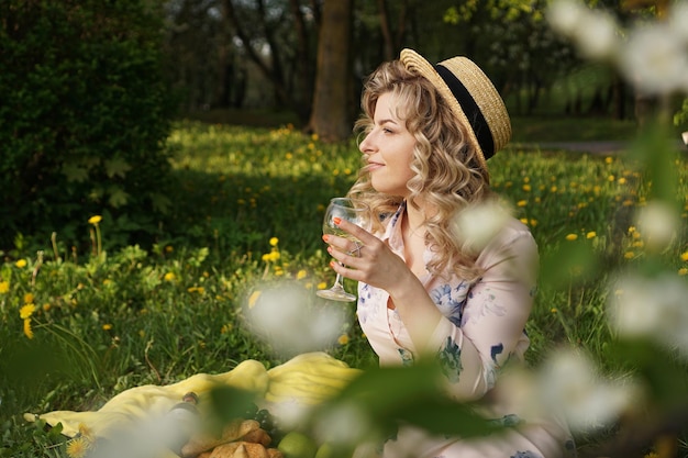 麦わら帽子のブロンドの髪を持つ美しい若い女性は、夏のピクニック中にワインを飲み、庭の格子縞に座っています