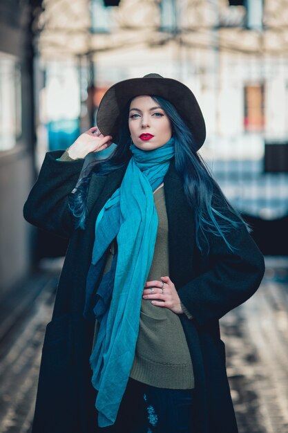Beautiful young woman with blue hair in nice black coat, jeans and hat.