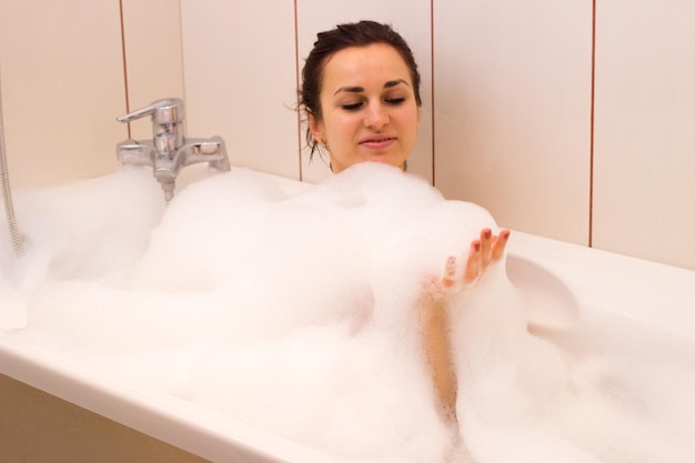 Beautiful young woman with bundle on her head lying in the tub full of foam in her bathroom