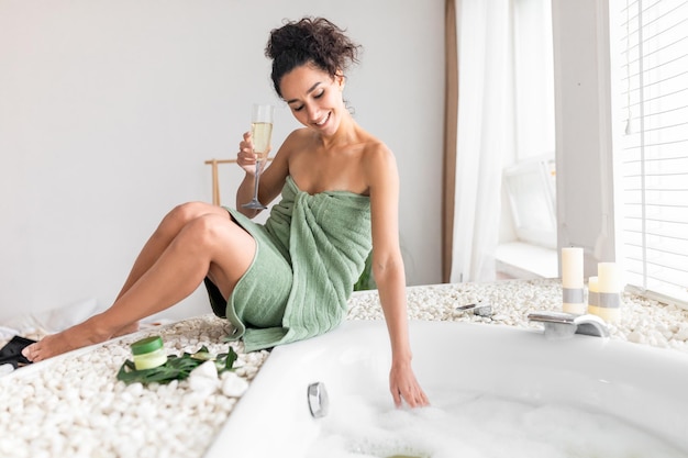 Beautiful young woman with champagne preparing bubble bath checking water temperature at home free