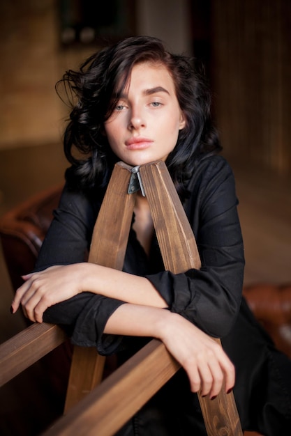 Beautiful young woman with curly dark hair and natural makeup sitting on a dark leather chairs