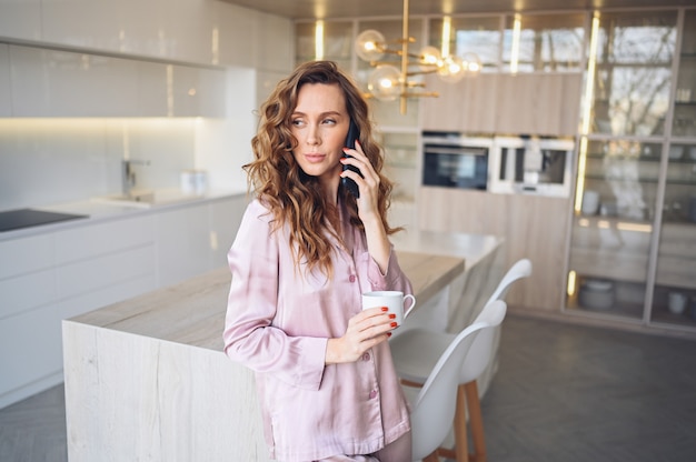 Beautiful young woman with curly hair drinking coffee at home in lazy weekend morning in cozy pink pajamas.