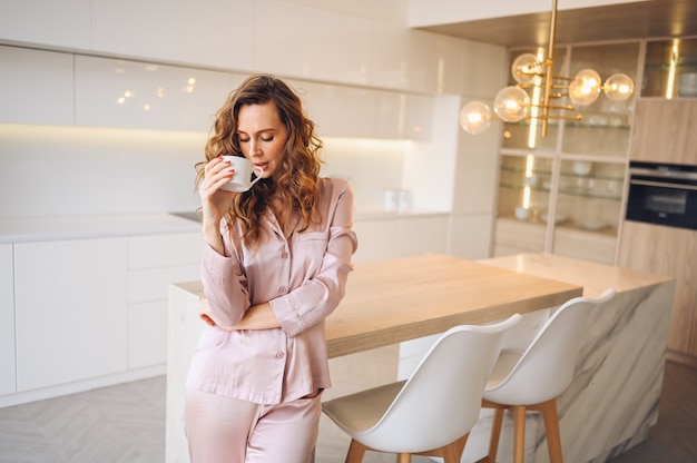 Beautiful young woman with curly hair drinking coffee in weekend morning in cozy pink pajamas. Lady in scandinavian style white kitchen modern interior.