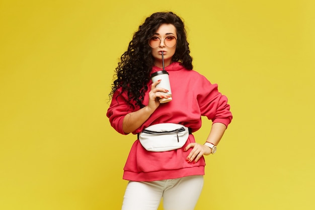 Beautiful young woman with curly hair in pink hoodie and with belt-bagh posing with a coffee cup to go, isolated on the yellow