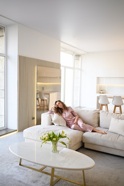 Beautiful young woman with curly hair in  pink pajamas lying at white couch sofa in the morning. Scandinavian style living room & kitchen interior. 