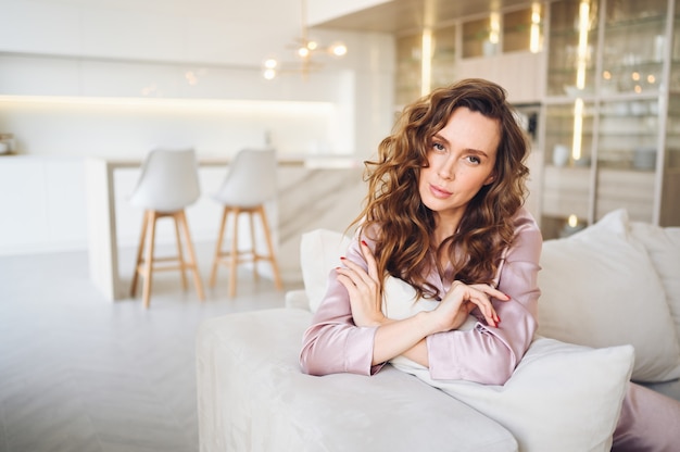 Beautiful young woman with curly hair in  pink pajamas sitting at white couch or sofa in the morning. Scandinavian style living room & kitchen interior. 