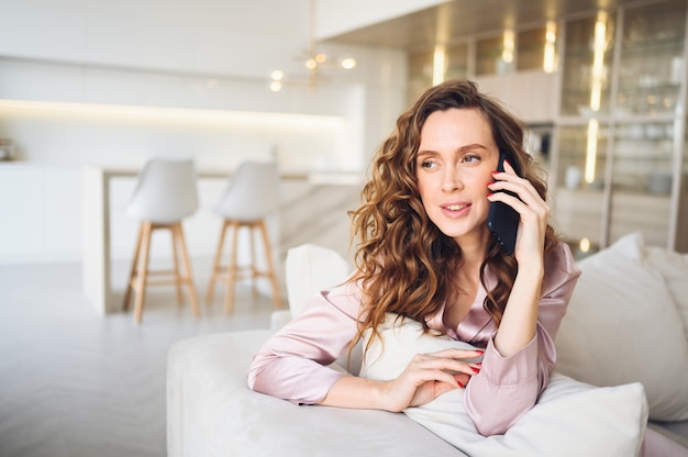 Beautiful young woman with curly hair in  pink pajamas at white couch in the morning. Lady speaking phone in scandinavian style living room interior.