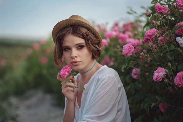 Beautiful young woman with curly hair posing near roses in a garden The concept of perfume advertising