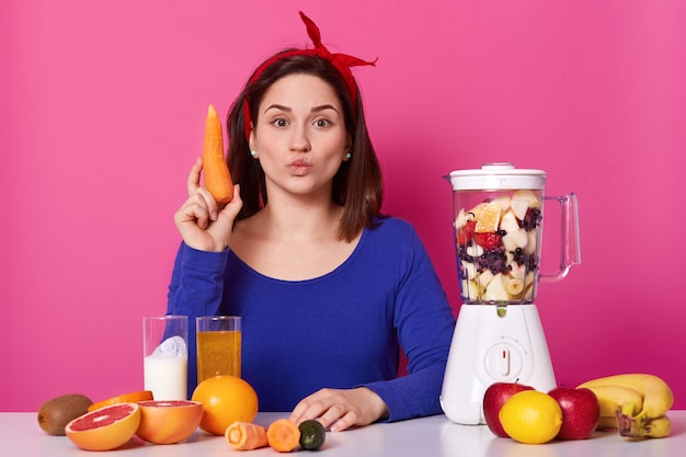 Beautiful young woman with dark hair, keeps lips rounded, holds carrot in hands, makes fresh smoothies, has blender filled with fruit on table. Female likes healthy eating. Lifestyle concept.