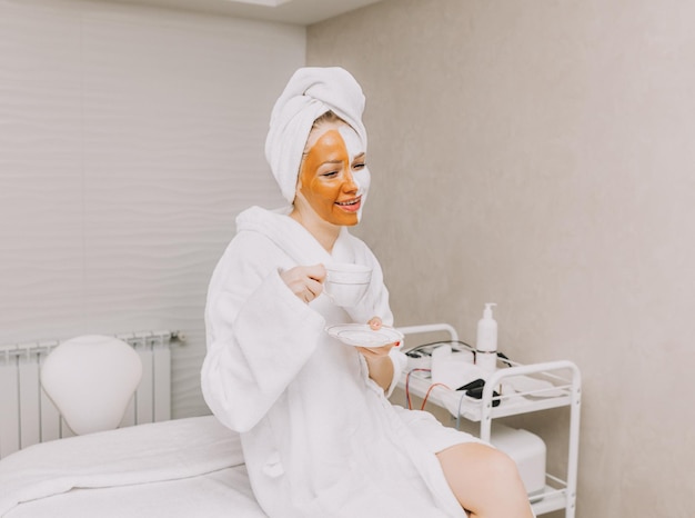 Beautiful young woman with a face mask drinks coffee in the beauticians office