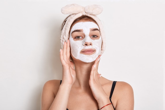 Beautiful young woman with facial mask, beauty treatment isolated, keeping palms on cheeks, looks confident and satisfied.