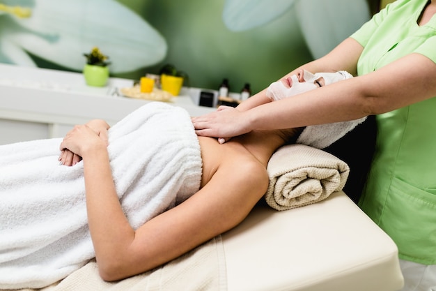 Beautiful young woman with facial mask getting spa massage treatment at beauty spa salon. Facial beauty treatment.