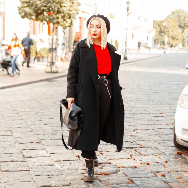 Beautiful young woman with a fashion bag in a black coat with a beret walks in the autumn day in the city