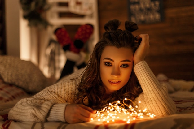 Bella giovane donna con luci festive in un maglione lavorato a maglia vintage che riposa su un letto alla vigilia di natale