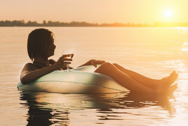 Photo beautiful young woman with a glass of wine on inflatable ring in water during sunset