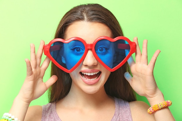 Beautiful young woman with heartshaped glasses on color background