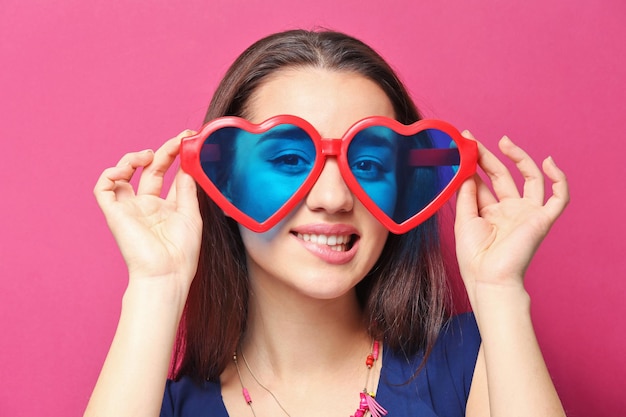 Bella giovane donna con gli occhiali a forma di cuore su sfondo colorato