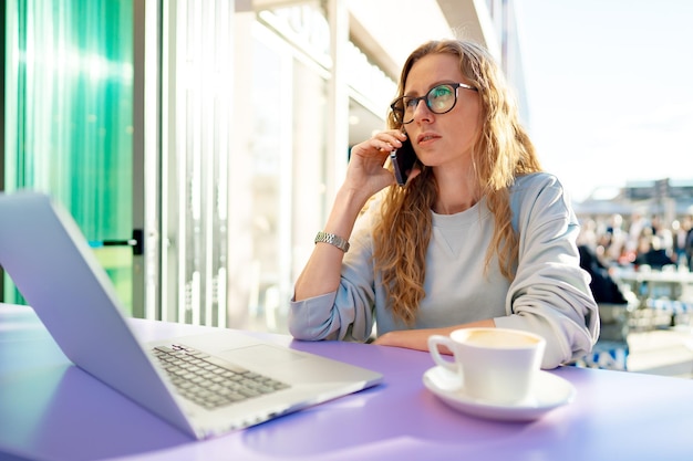 Bella giovane donna con il computer portatile che parla al telefono al caffè all'aperto