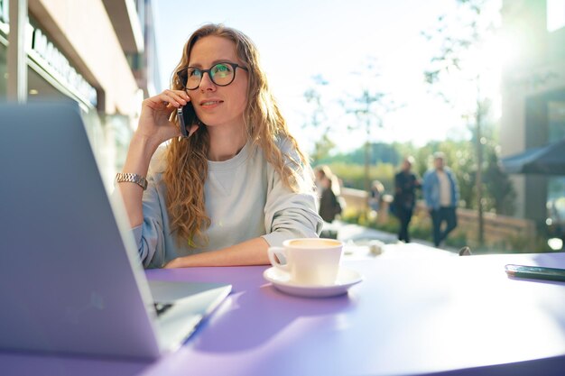 Bella giovane donna con il computer portatile che parla al telefono al caffè all'aperto