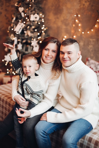 Beautiful young woman with long black wavy hair in sweater and trousers, attractive strong man with short dark hair in sweater and jeans spend Christmas time together with their son