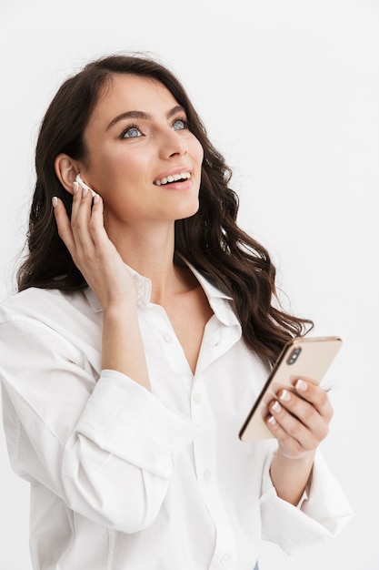 Beautiful young woman with long curly brunette hair wearing white shirt standing isolated over white wall, enjoying listening to music with earphones, holding mobile phone