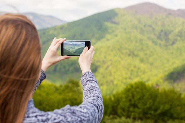 長い髪の美しい若い女性は、緑の自然の彼女の黒い電話で写真を撮っています