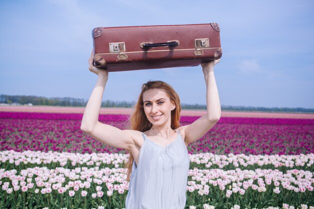 Beautiful young woman with long red hair wearing in white dress standing with luggage on colorful tulip field.