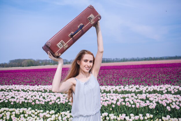 Foto bella giovane donna con lunghi capelli rossi che indossa in abito bianco in piedi con i bagagli sul campo di tulipani colorati.