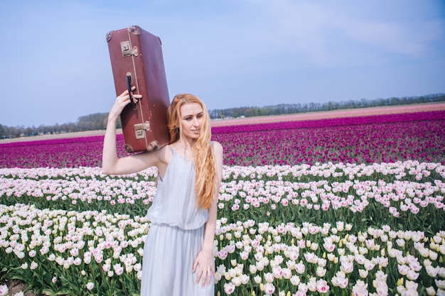 Beautiful young woman with long red hair wearing in white dress standing with luggage on colorful tulip field.