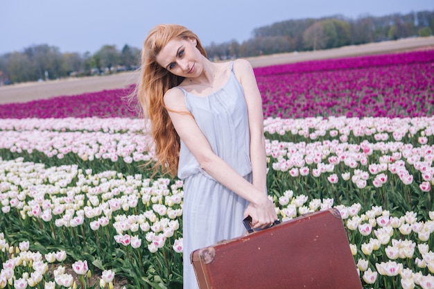 Beautiful young woman with long red hair wearing in white dress standing with old vintage suitcase on colorful tulip field.