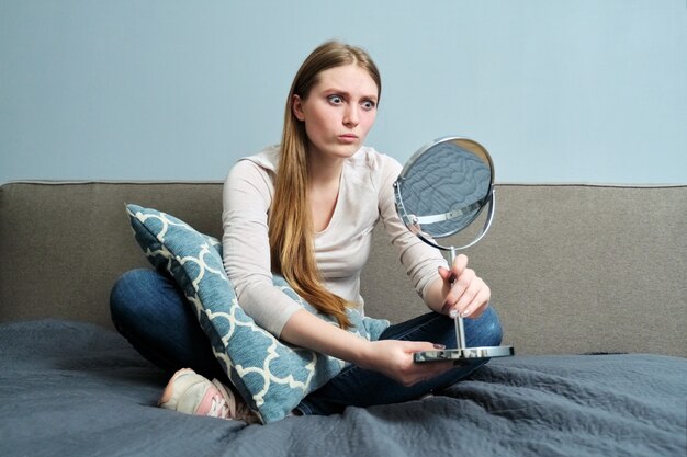 Beautiful young woman with make-up mirror sitting at home in bed, girl makes emotions, looks at herself in mirror