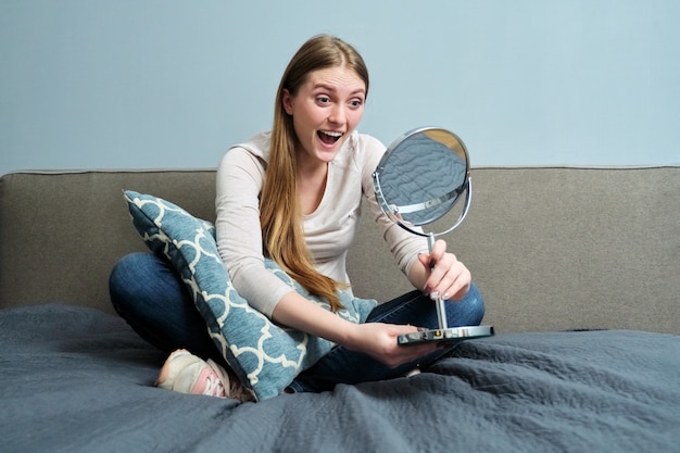 Beautiful young woman with make-up mirror sitting at home in bed, girl makes emotions, looks at herself in mirror