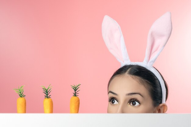 Beautiful young woman with pink bunny ears and toy carrot on pink.