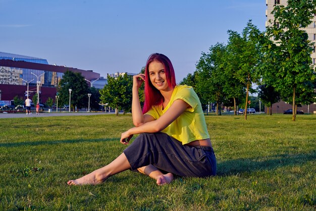 Beautiful young woman with pink hair is sitting on the lawn in a city park and smiling