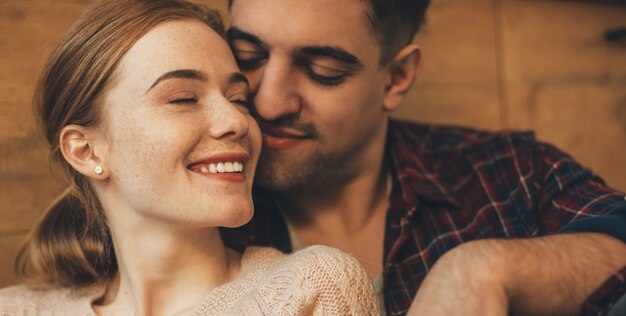 Beautiful young woman with read hair and freckles laughing with closed eyes while her boyfriend is embracing her from back sleeping with closed eyes on floor