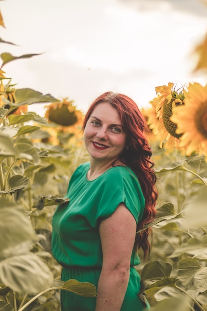 Beautiful young woman with red curly hair and a green dress posing in a sunflowers field