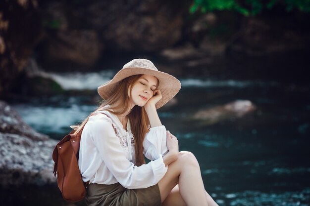 빨간 머리, 모자 및 숲의 시끄러운 강 근처 배낭과 함께 아름 다운 젊은 여자.