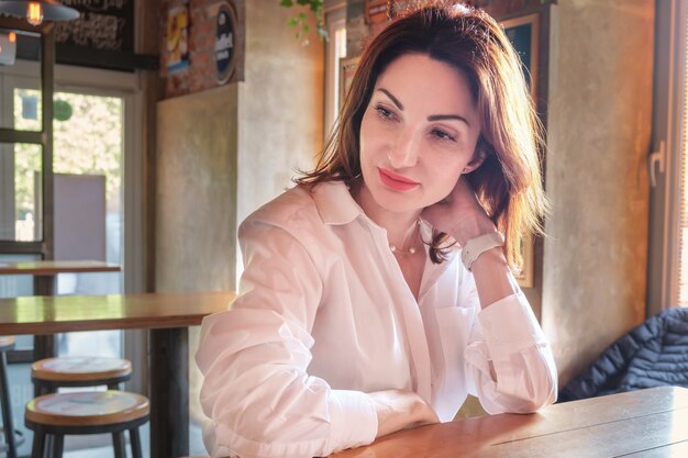 Фото Красивая молодая женщина с рыжими волосами в белой рубашке сидит за деревянным столом в баре и задумчивая. портрет молодой женщины. женщина ждет встречи.